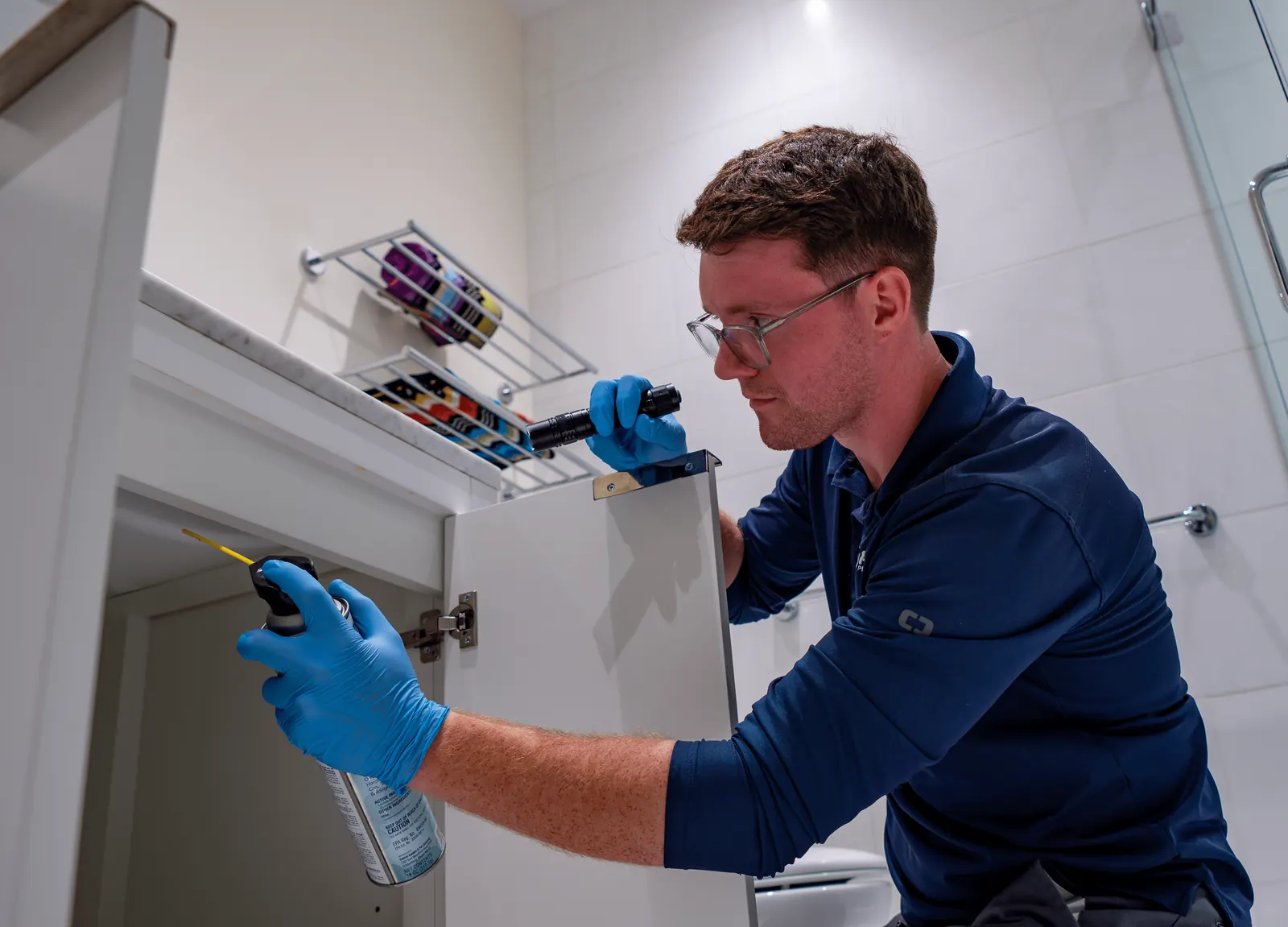 Prodigy technician inspecting under a bathroom sink cabinet with a flashlight and applying treatment